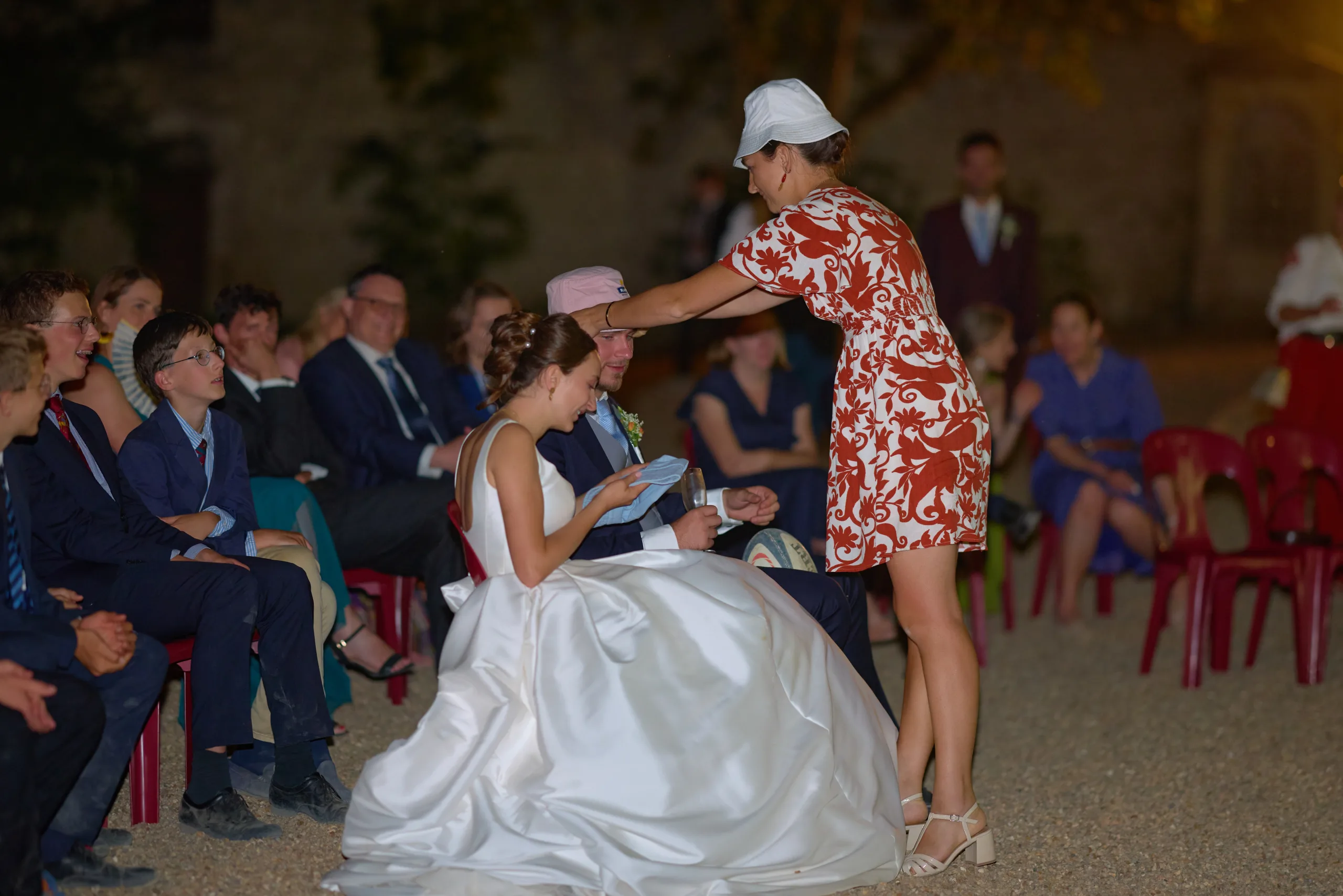 Une invitée au chapeau blanc se penche vers les mariés assis
